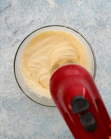 two hands beating cream cheese mixture in a glass bowl using a red electric beater.