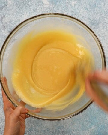 two hands whisking eggs in a large glass bowl.