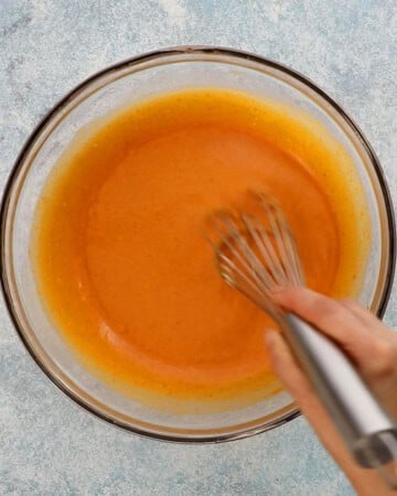 two hands whisking orange pumpkin mixture in a large glass bowl.