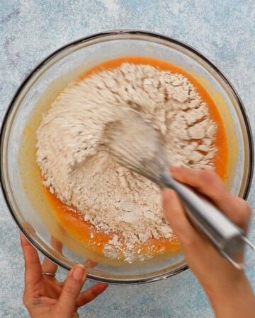 two hands mixing pumpkin cake batter in a large glass bowl.