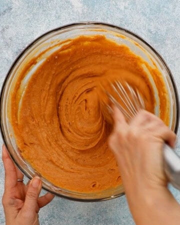 two hands mixing pumpkin cake batter in a large glass bowl.