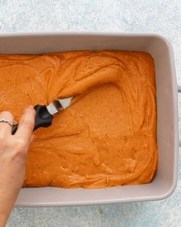 a hand spreading orange pumpkin cake batter in a grey pan using a flat spatula. 