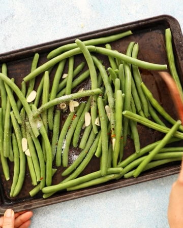 two hands stirring whole green beans along with sliced garlic in a black baking sheet.