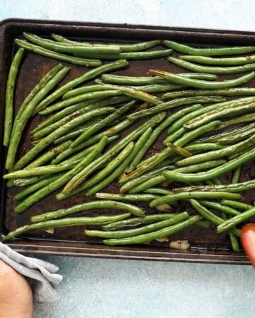 two hands tossing roasted green beans in a black baking sheet.