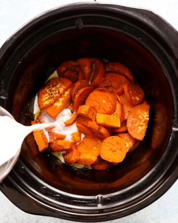 a hand pouring white milk into a black pot filled with cooked sweet potatoes.