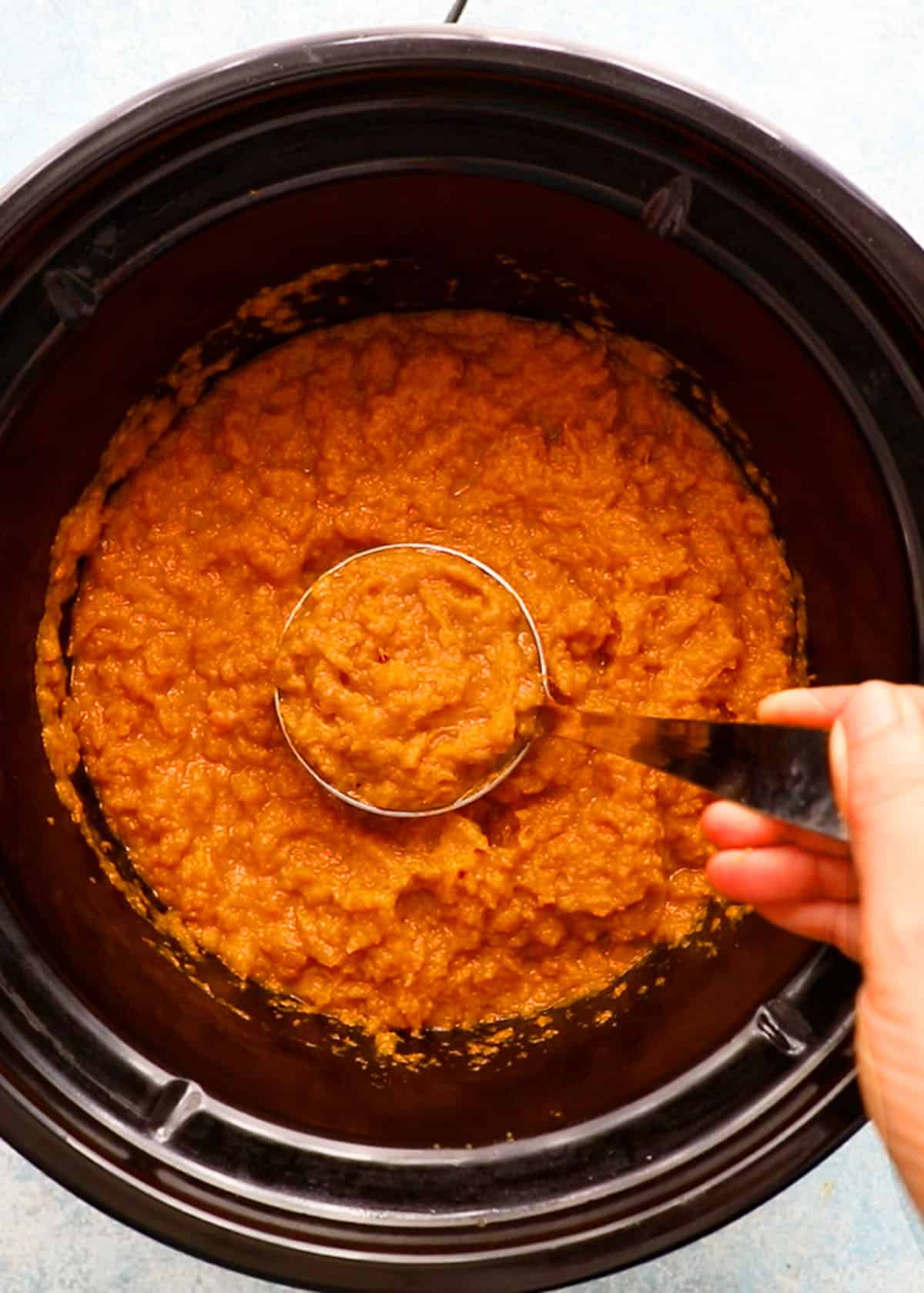 a hand holding one ladle full of mashed sweet potatoes in a black pot.